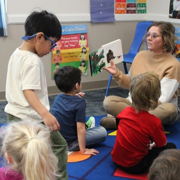 A teacher reads an illustrated picture book to a small group of preschool children gathered on a classroom rug.