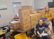 Three ESU 13 staff members organize family literacy kits among stacks of cardboard boxes.