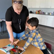 An educator and a young student explore an open picture book together, pointing to illustrations on the pages.