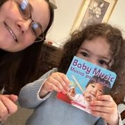 A young child holds up a bilingual board book titled Baby Music / Música para bebés while sitting beside an adult.
