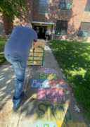 A staff member pushes a cart stacked with boxes toward a school entrance along a colorful sidewalk.
